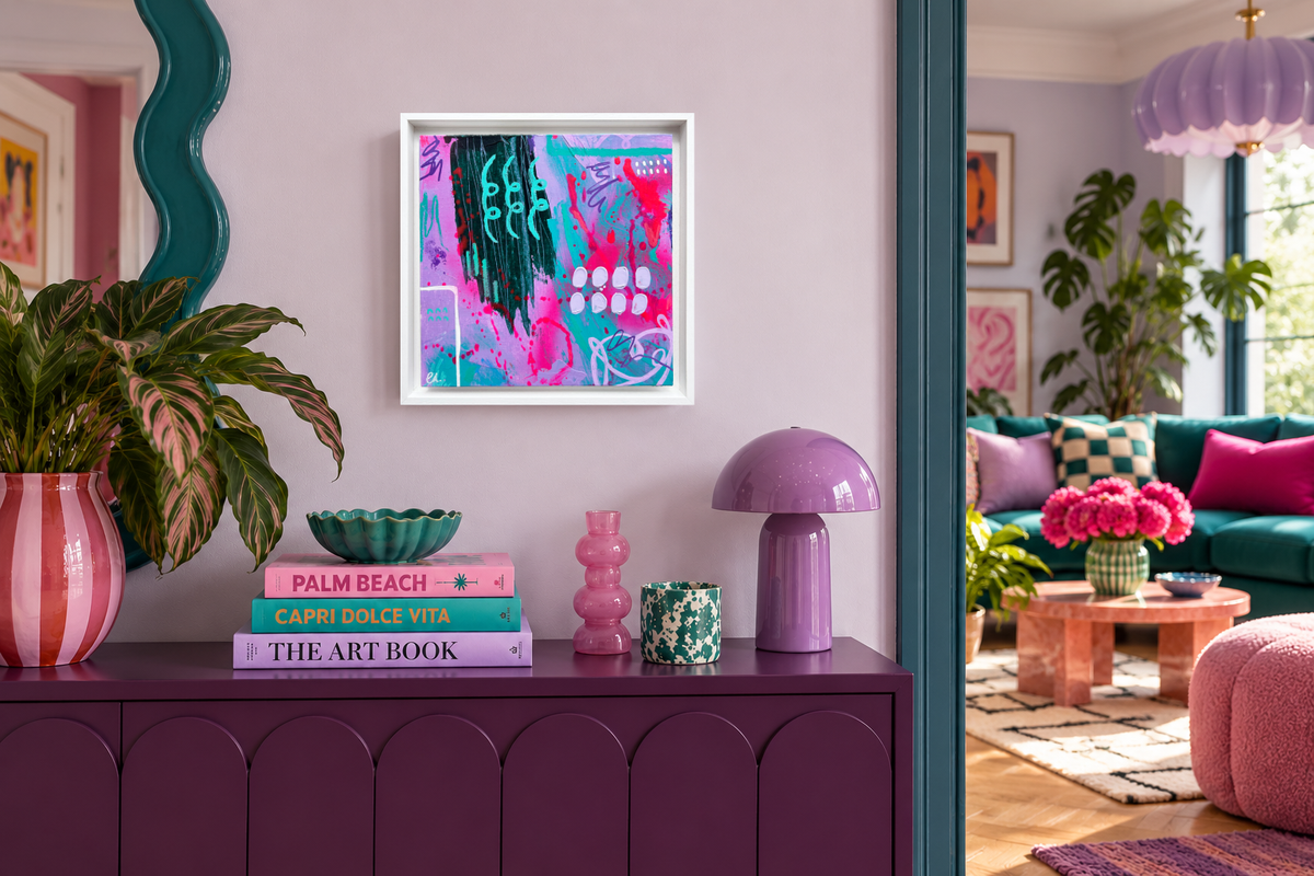 A framed abstract art print with vibrant pink, teal, and purple brushstrokes hangs on a lavender wall above a purple console table in a styled living room.