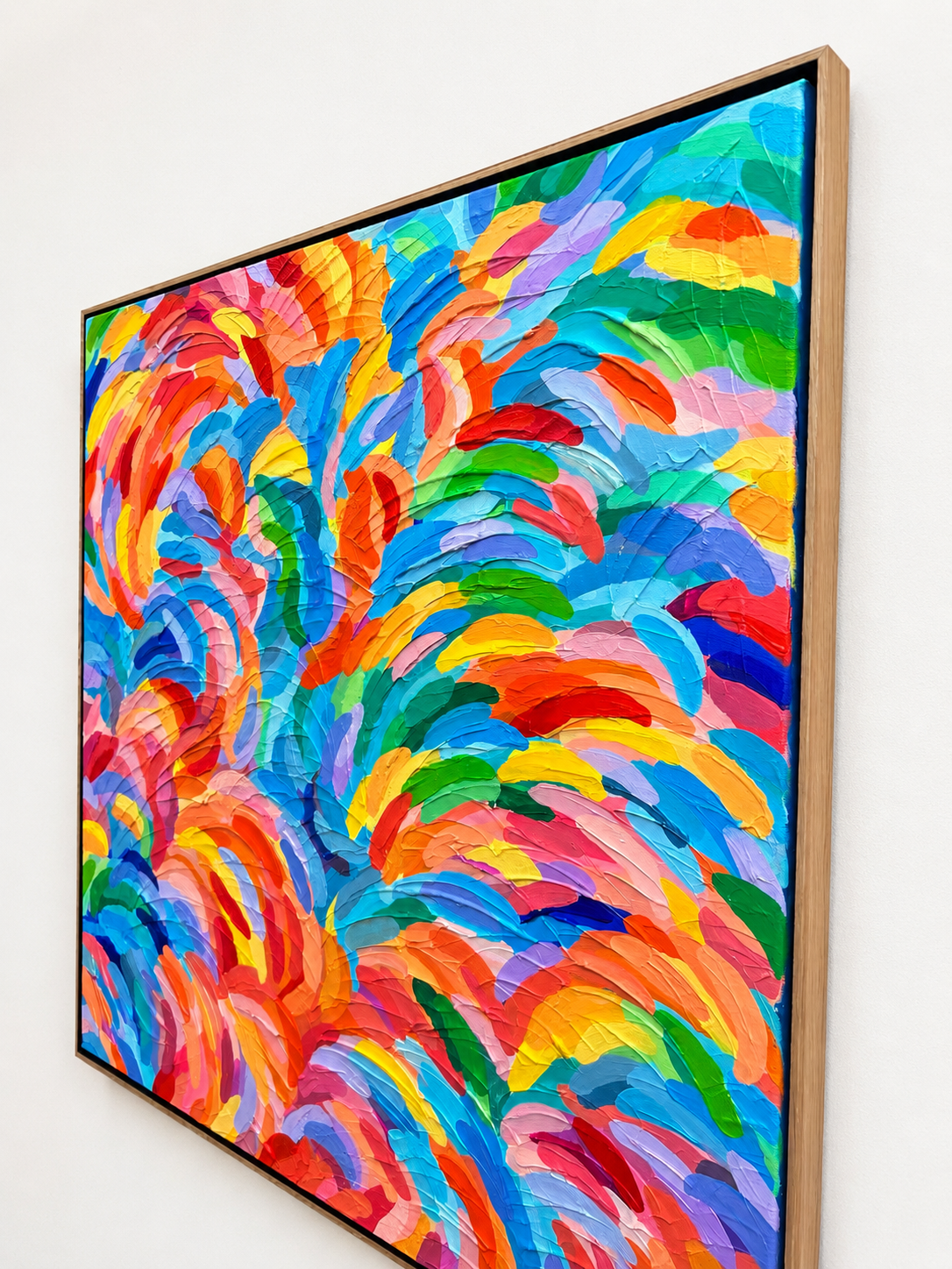A framed abstract canvas painting featuring a vibrant, textured impasto brushstroke pattern in a rainbow of colors, hung on a plain white wall.