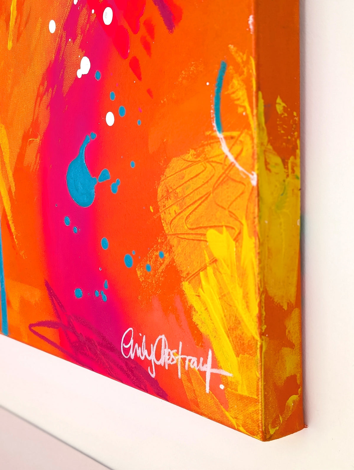 Close-up of a colorful abstract canvas painting showing bright orange, pink, yellow, and blue brushstrokes with a white signature near the bottom, mounted against a white wall.
